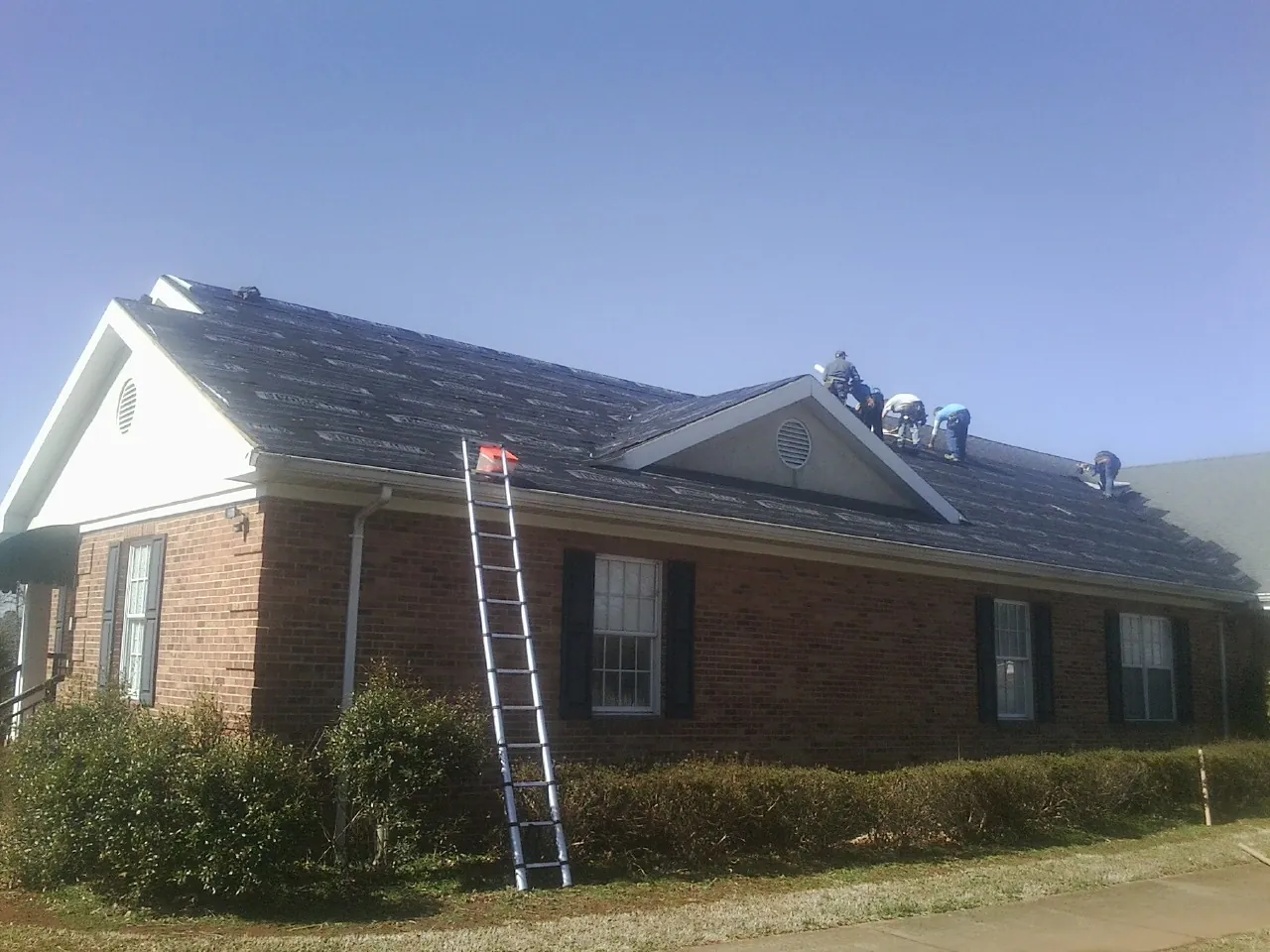 Crew tearing shingles off a brick ranch roof with a ladder set — Metal Roof Repair work by our crew in Yelm