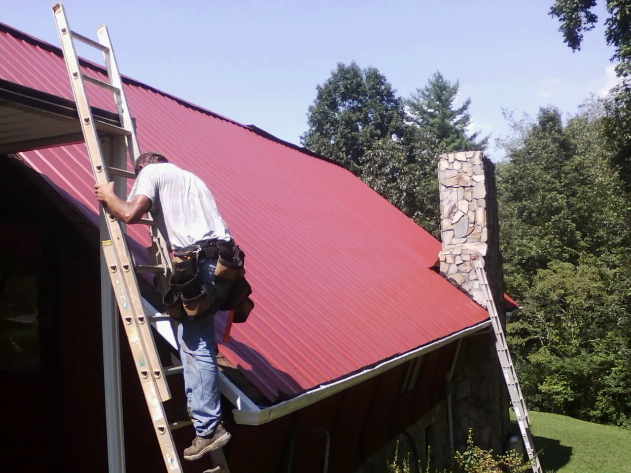 Roofer on a ladder beside a red metal roof and stone chimney — Soffit & Fascia Repair by Roofing Brothers in Yelm